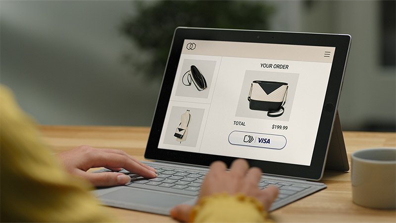 A person in front of a laptop paying for a bag in an online shop using Visa's click-to-pay payment options.